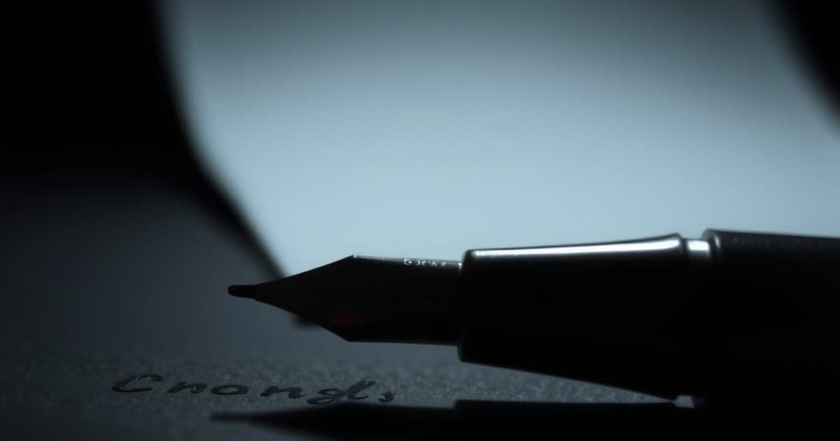 Ponta metálica de uma caneta-tinteiro traçando uma linha de tinta sobre papel escuro, em foto de luz lateral.