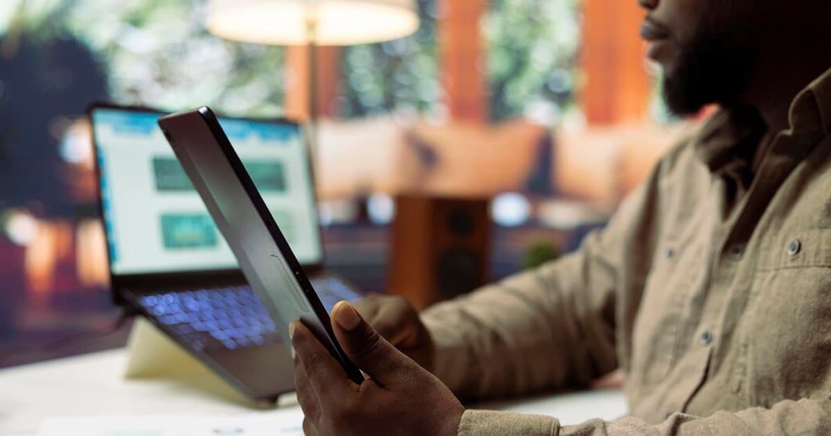 Pessoa segurando um tablet em ambiente de escritório com laptop desfocado ao fundo.