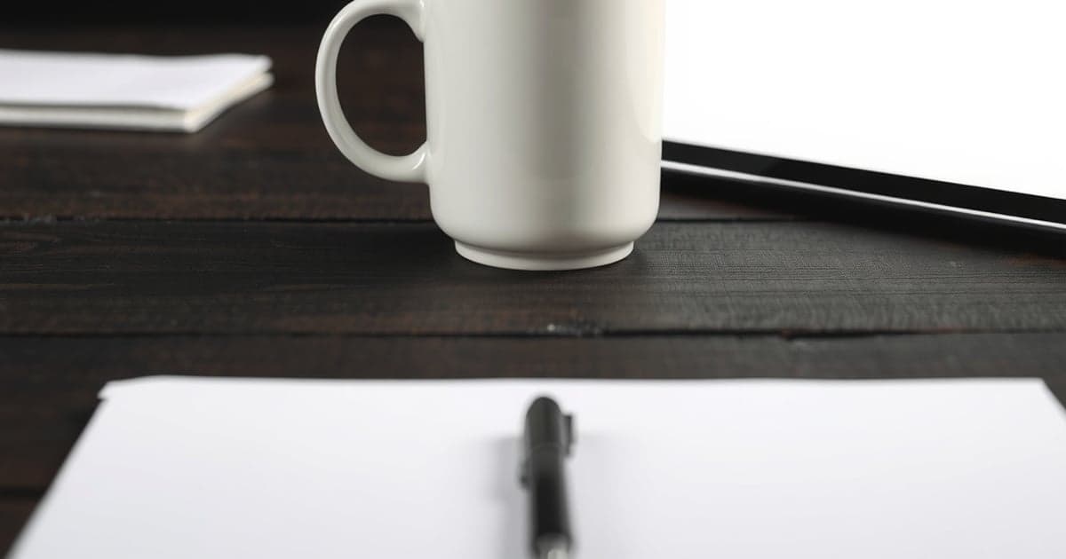Caneca branca de cerâmica sobre uma mesa de madeira escura, ao lado de uma folha em branco e uma caneta, em luz suave.