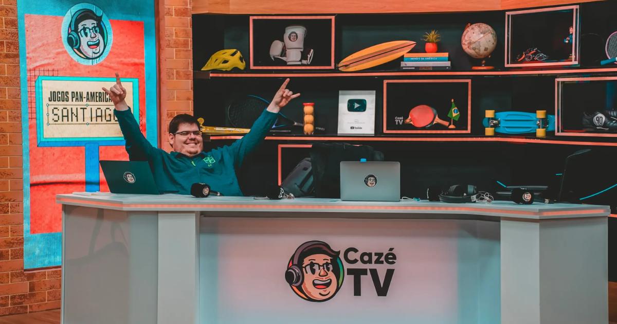 Apresentador comemorando com os braços erguidos na bancada do estúdio da CazéTV