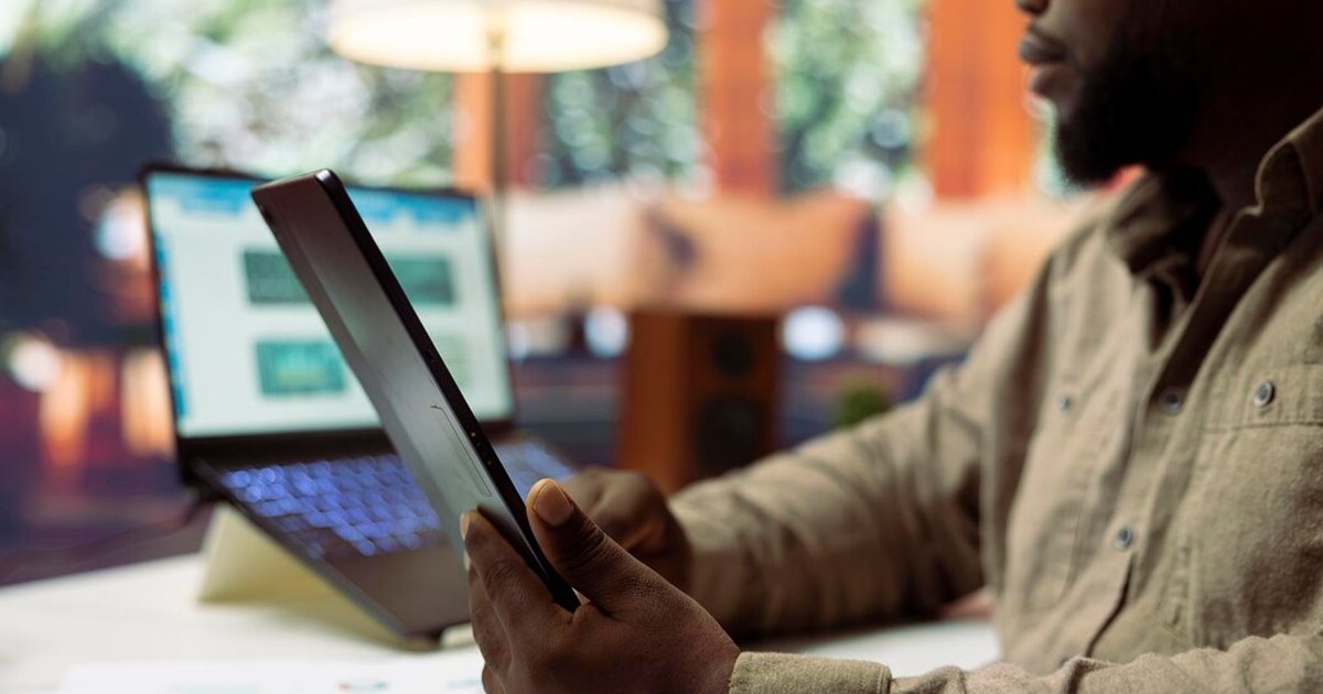 Pessoa segurando um tablet em ambiente de escritório com laptop desfocado ao fundo.