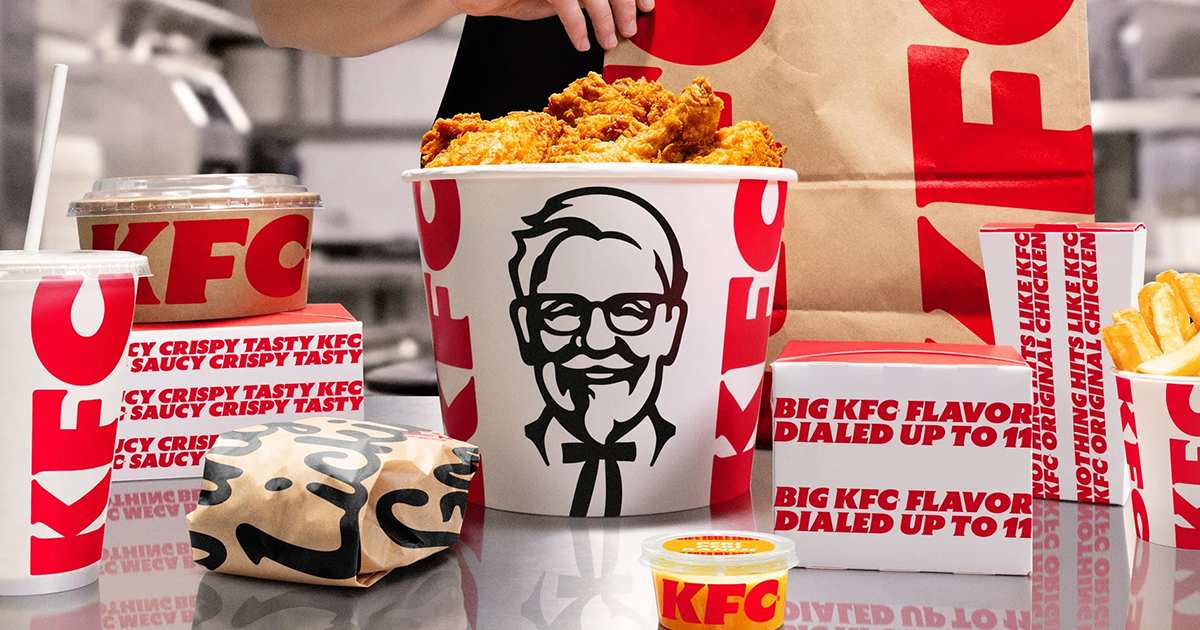 Balde de frango do KFC com o logo do Coronel Sanders, cercado de embalagens e sacola vermelha da marca