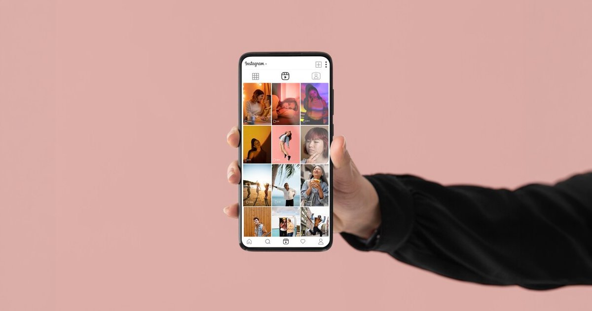 Mão segurando um celular exibindo o feed do Instagram, fundo rosa liso.